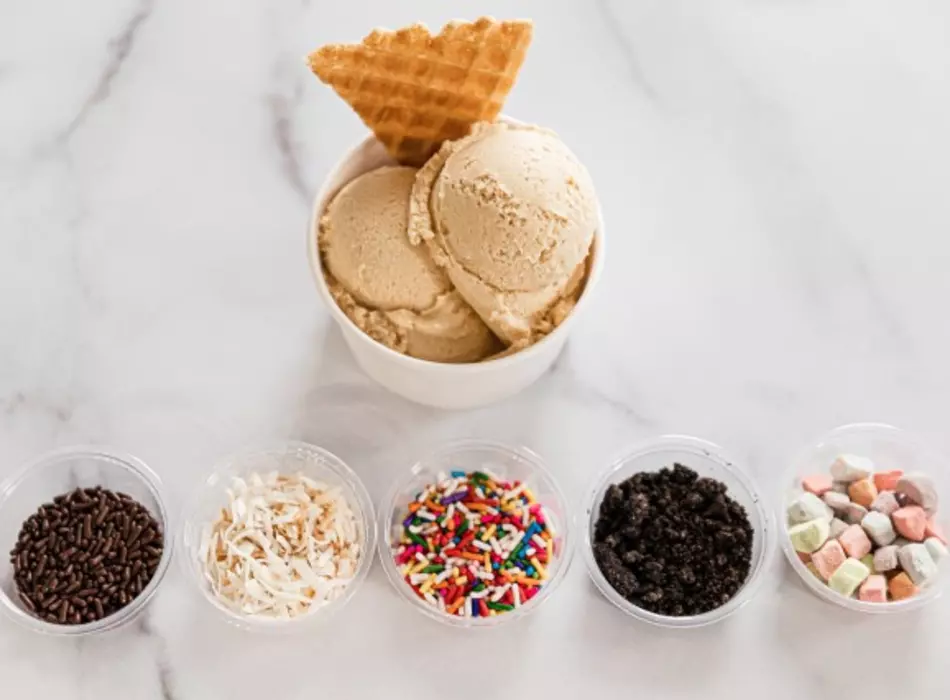 gelato scoops in a cup with a waffle wedge and 5 containers of toppings lined up below it